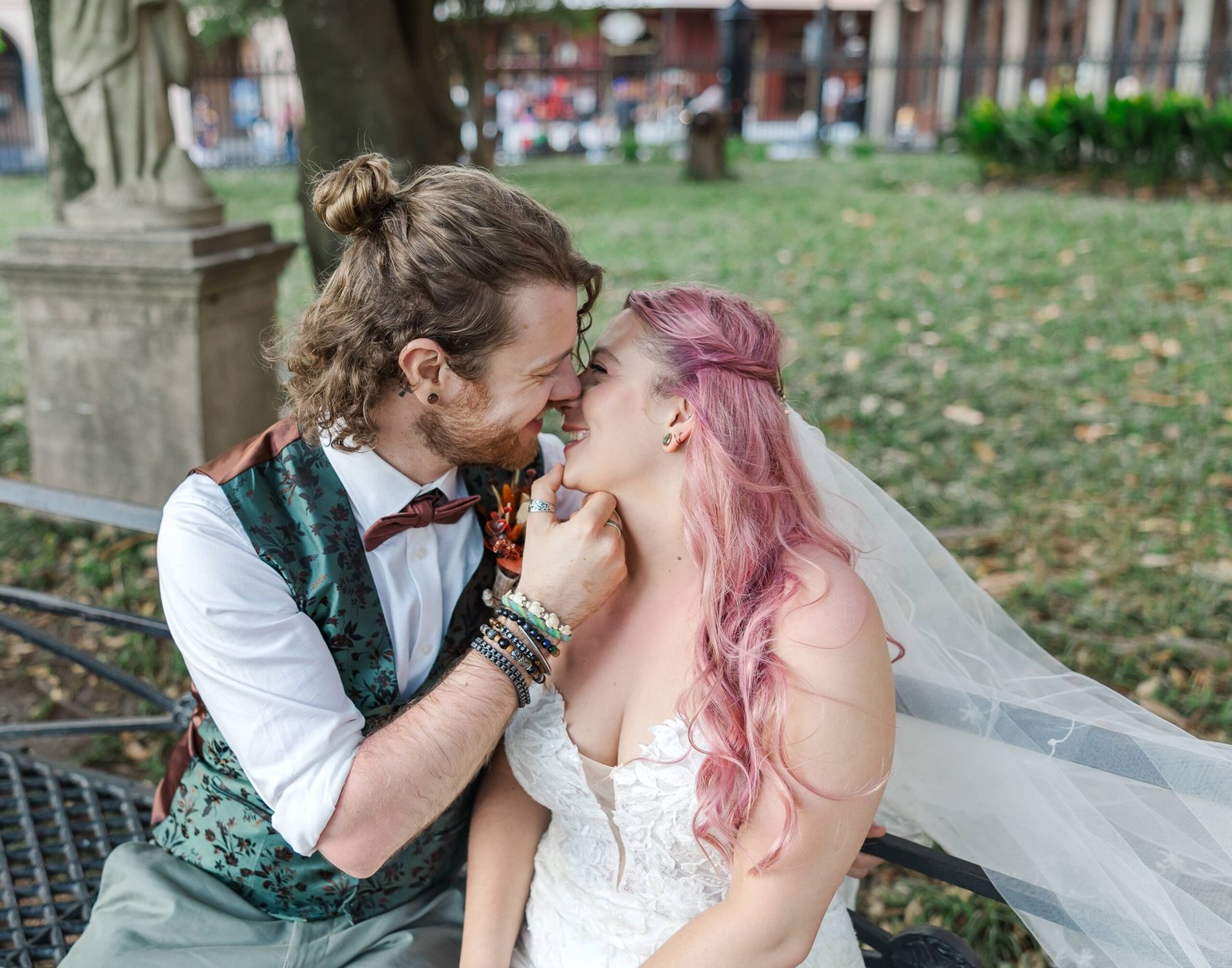 A joyful bride with pink hair and a groom with a man bun share a tender kiss in a lush park, surrounded by greenery, captured during their New Orleans wedding elopement.