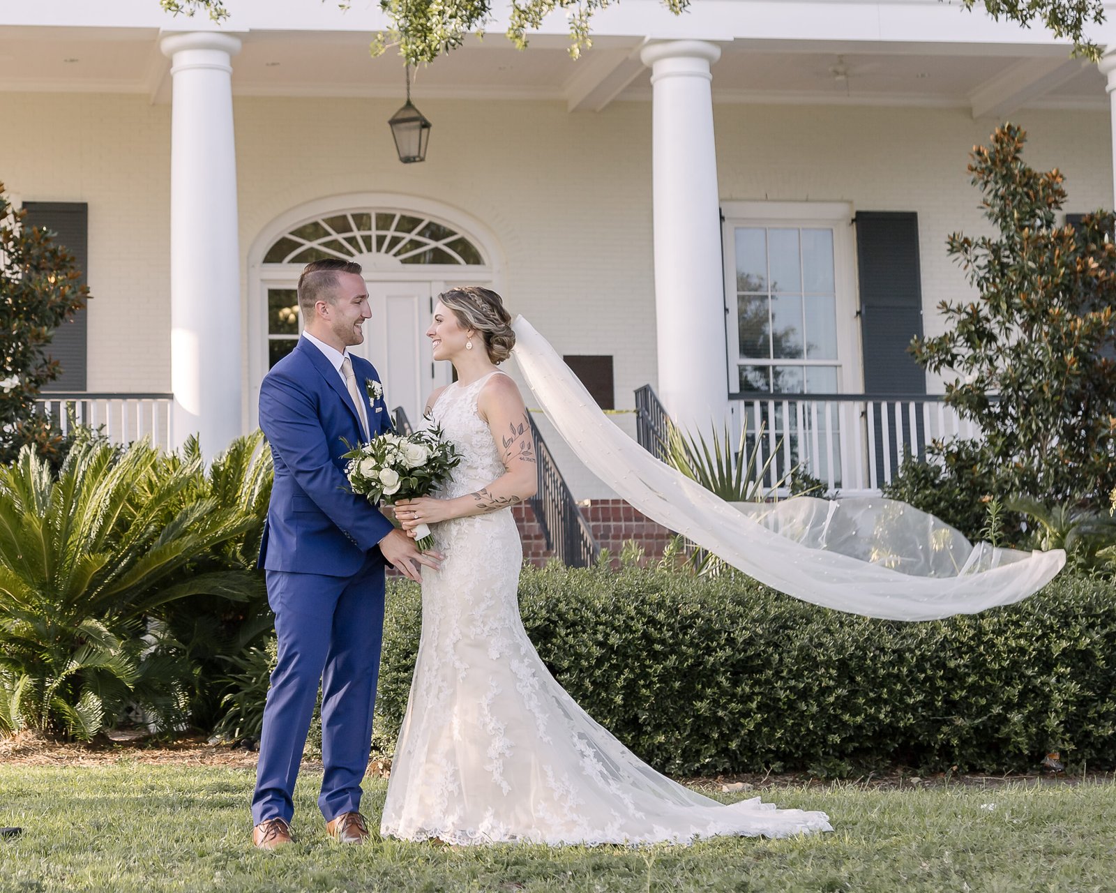 Couple who eloped smiling at each other in front of a biloxi visitors center in Biloxi, Mississippi, with the bride’s veil flowing dramatically in the breeze.