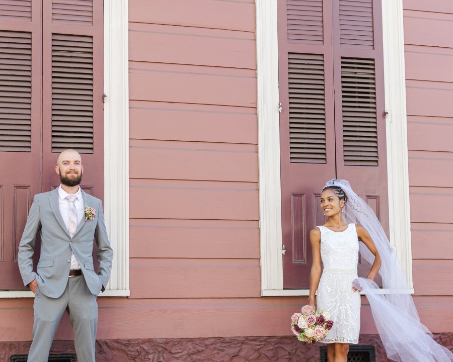 Newly married couple smiling at each other in front of a colorful turquoise and blue New Orleans shotgun house.